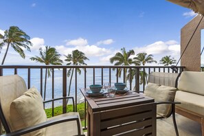 A balcony with a view of the ocean, palm trees, and a table set with blue bowls and glasses. - Oceanfront Maui condo with whale watching views, hot tub summer deals (Wailuku)