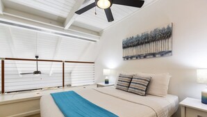 Une chambre avec un grand lit, un ventilateur de plafond, une fenêtre avec des stores, une table de chevet avec une lampe et un tableau au mur.