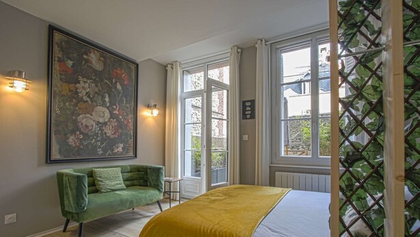 Une chambre avec un canapé vert, un grand tableau floral et un lit recouvert d’une couverture jaune.