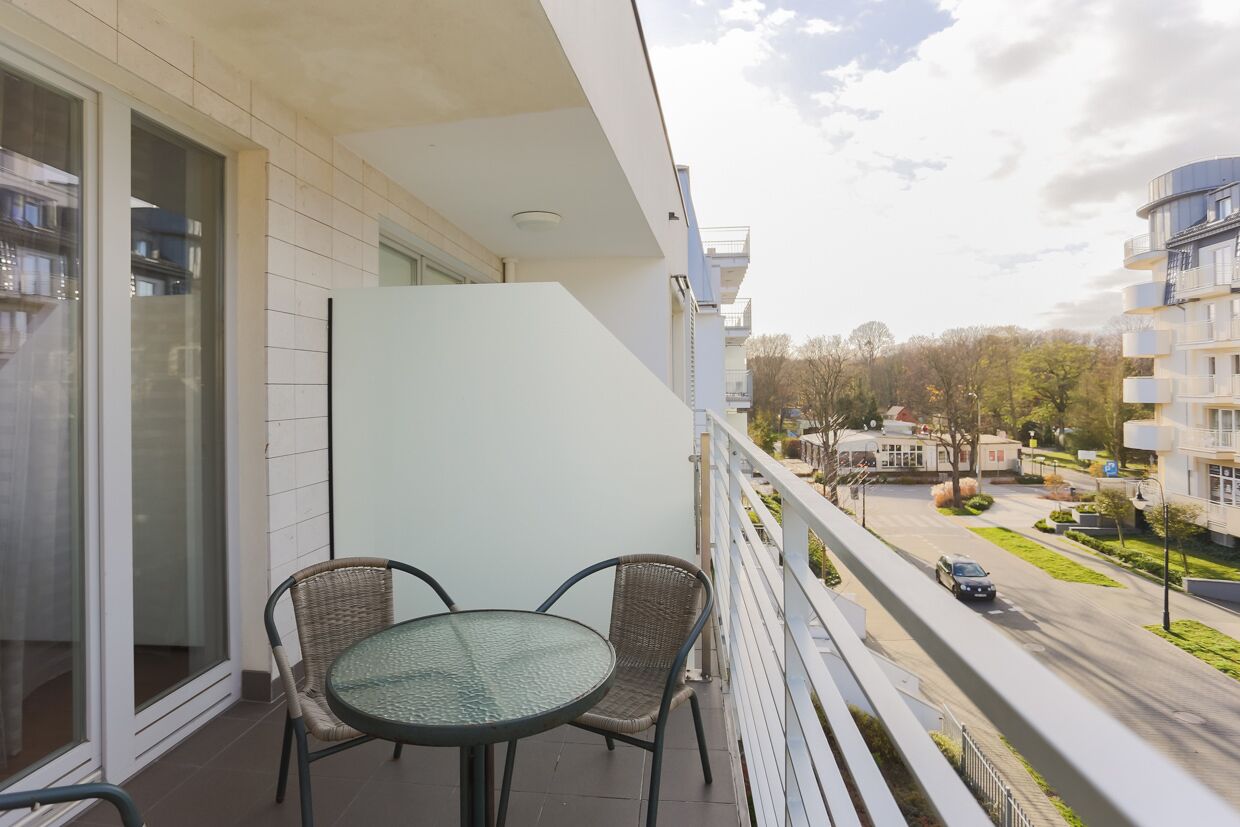 Un balcón con una mesa de vidrio y sillas de mimbre, con vista a una calle con edificios y árboles.