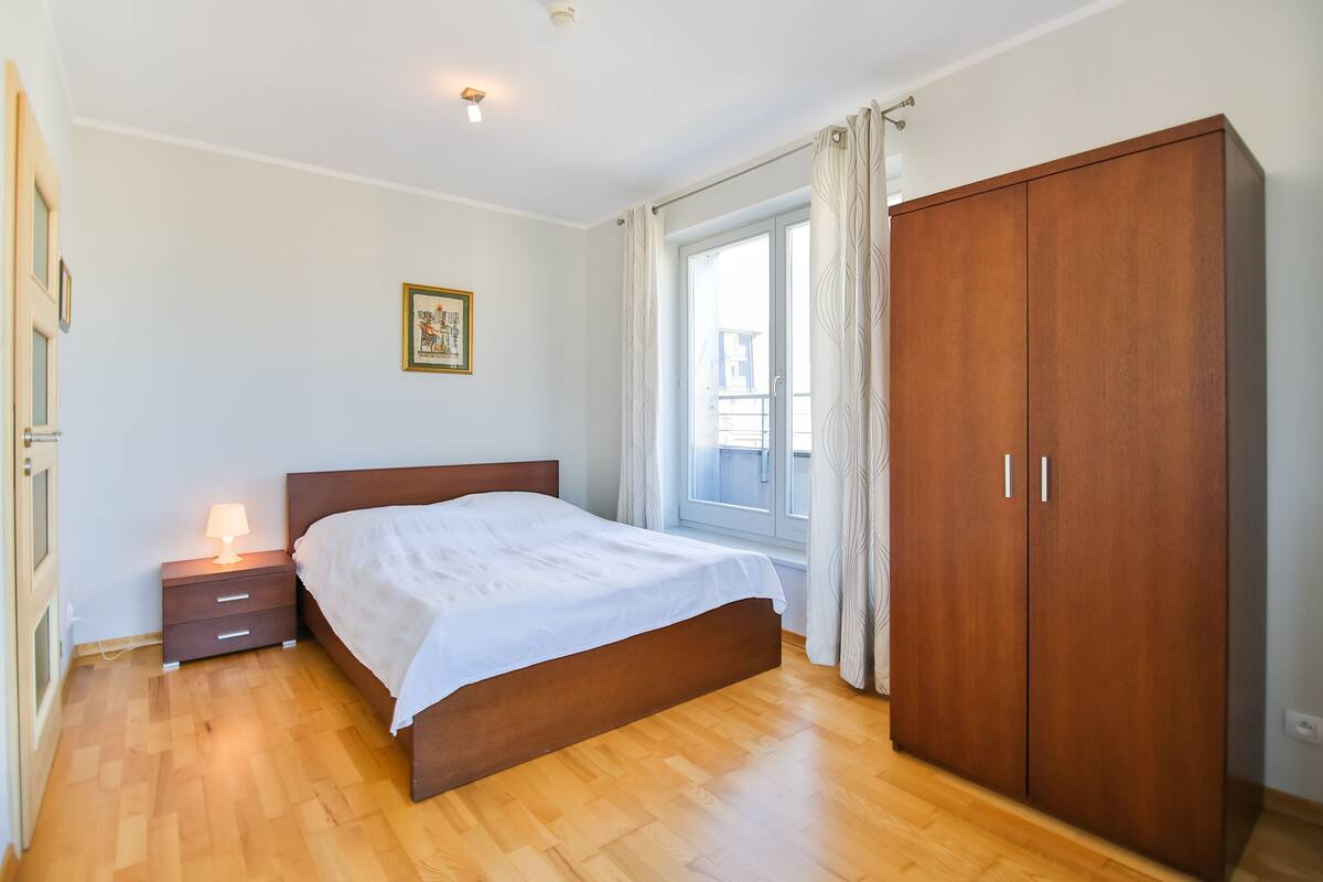 Een slaapkamer met een houten bed, een nachtkastje met een lamp, een kledingkast en een raam met gordijnen.
