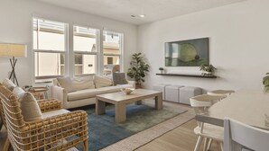Un salón amplio con sofá, una mesita de centro y sillones de mimbre. Se observan ventanas grandes, un cuadro en la pared y una planta.