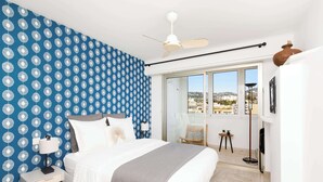 A bedroom with a bed, a nightstand, a lamp, a ceiling fan, a window with a view, and a small table with a plant. - On la Croisette XXL 1 bedroom with amazing sea view (Cannes)