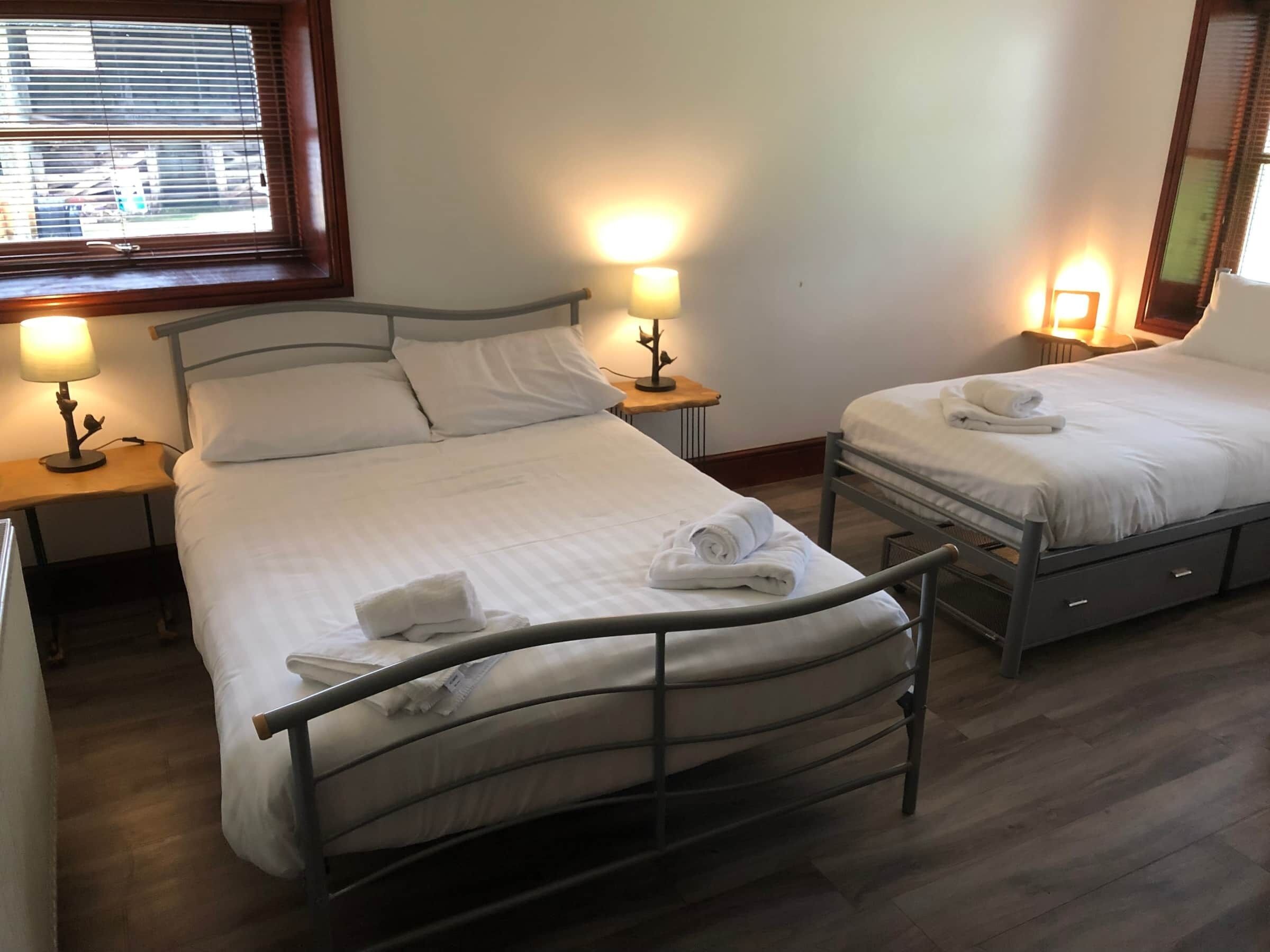 A hotel room with two beds, each with a nightstand and lamp. There is a window with blinds, and the room has a wooden floor.