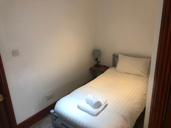 A small, single-bed room with a wooden headboard, a bedside table with a lamp, and a folded towel on the bed.