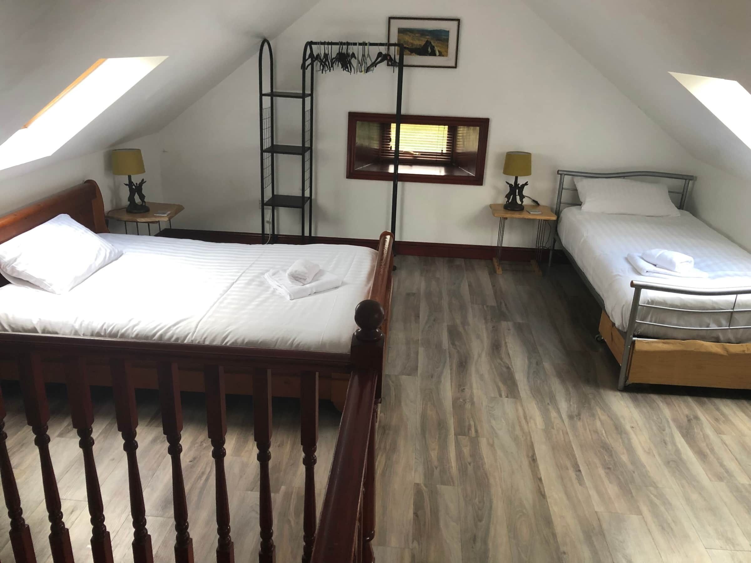Ein Zimmer mit zwei Betten, Holzboden, einer Himmelsleiter, einem kleinen Tisch mit Lampe und einem Fenster mit Jalousien.