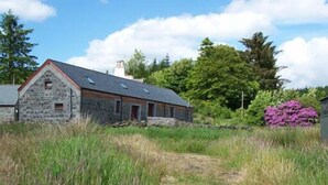 A stone building with a sloped roof, surrounded by tall grass and trees. - Maberry Cottage, Waterside Newton Stewart. (Polbae)