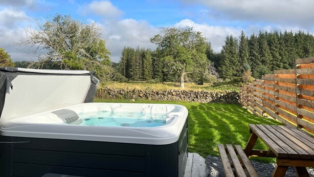 A hot tub in a backyard with a stone wall and wooden fence, surrounded by trees and a grassy lawn. - Maberry Cottage, Waterside Newton Stewart. (Polbae)