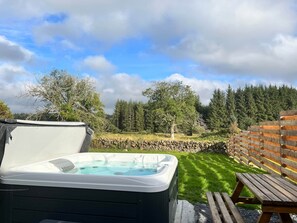 A hot tub in a backyard with a stone wall and wooden fence, surrounded by trees and a grassy lawn. - Maberry Cottage, Waterside Newton Stewart. (Polbae)