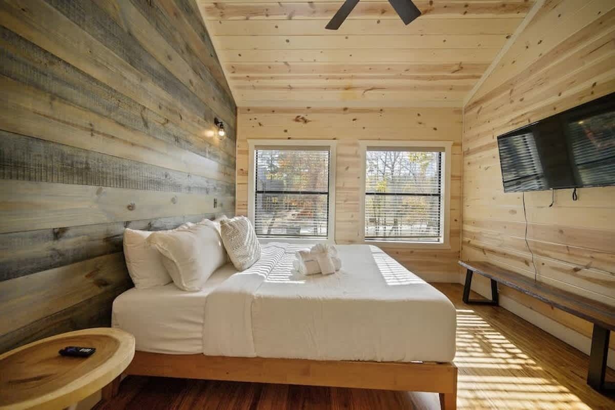Un dormitorio de cabaña de madera con una cama, dos ventanas, un televisor y un banco de madera.