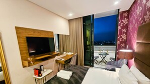 A hotel room with a large bed, a TV mounted on a wooden panel, a small table with a chair, a balcony with a table and chairs, and a view of the city at night. - Trendy Studio Apartment Centrally Located (Johannesburg)