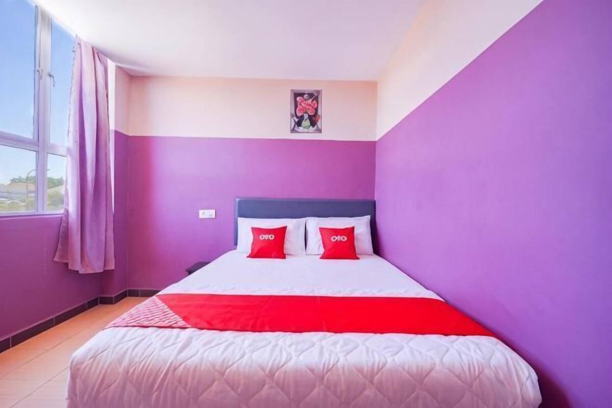 Ein Zimmer mit einem Bett, violetten Wänden, einem Fenster mit Vorhängen und einem gerahmten Bild an der Wand.