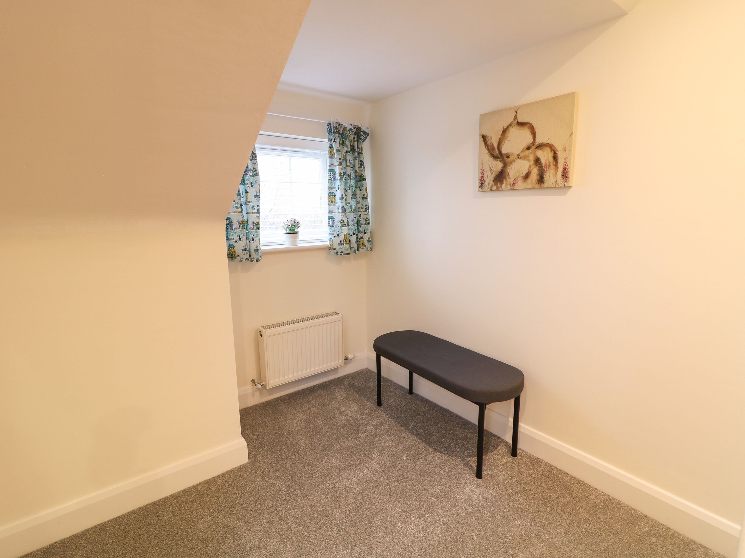 A small, carpeted room with a bench, a radiator, and a window with patterned curtains.