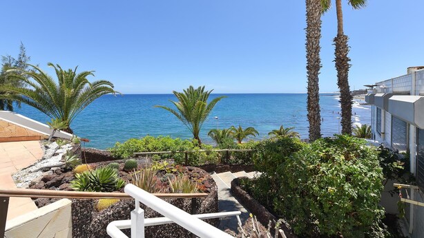Un balcón con vista al océano, palmeras y un jardín rocoso.