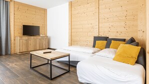 Une chambre moderne avec des murs lambrissés, un téléviseur à écran plat posé sur un meuble en bois, un lit avec une literie blanche et deux oreillers, une petite table en bois et un rideau gris.