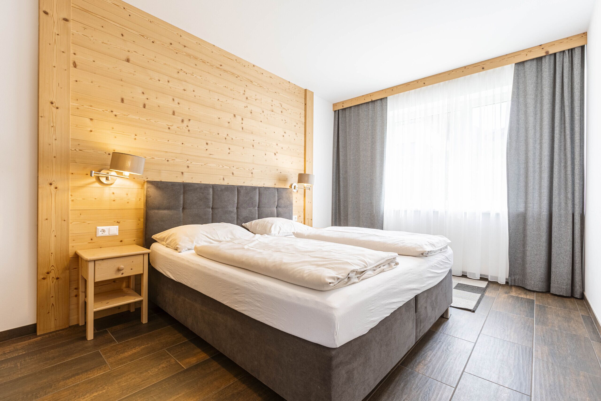 Una cama doble con cabecera gris, paredes de paneles de madera, dos lámparas de noche, una mesita de noche pequeña y una ventana grande con cortinas grises.