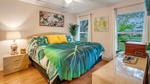 Une chambre à coucher comprenant un lit, une table de chevet, une lampe, un ventilateur de plafond et une fenêtre avec des rideaux.