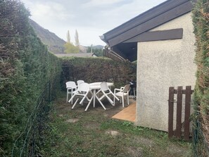 Un patio con sillas y mesa de plástico blanco, una parrilla y un pequeño edificio con techo inclinado.