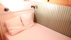 A small, single-bed room with a white bedspread and two pillows. There is a wooden headboard with a light switch and a small shelf above it. The wall has a vertical panel design.