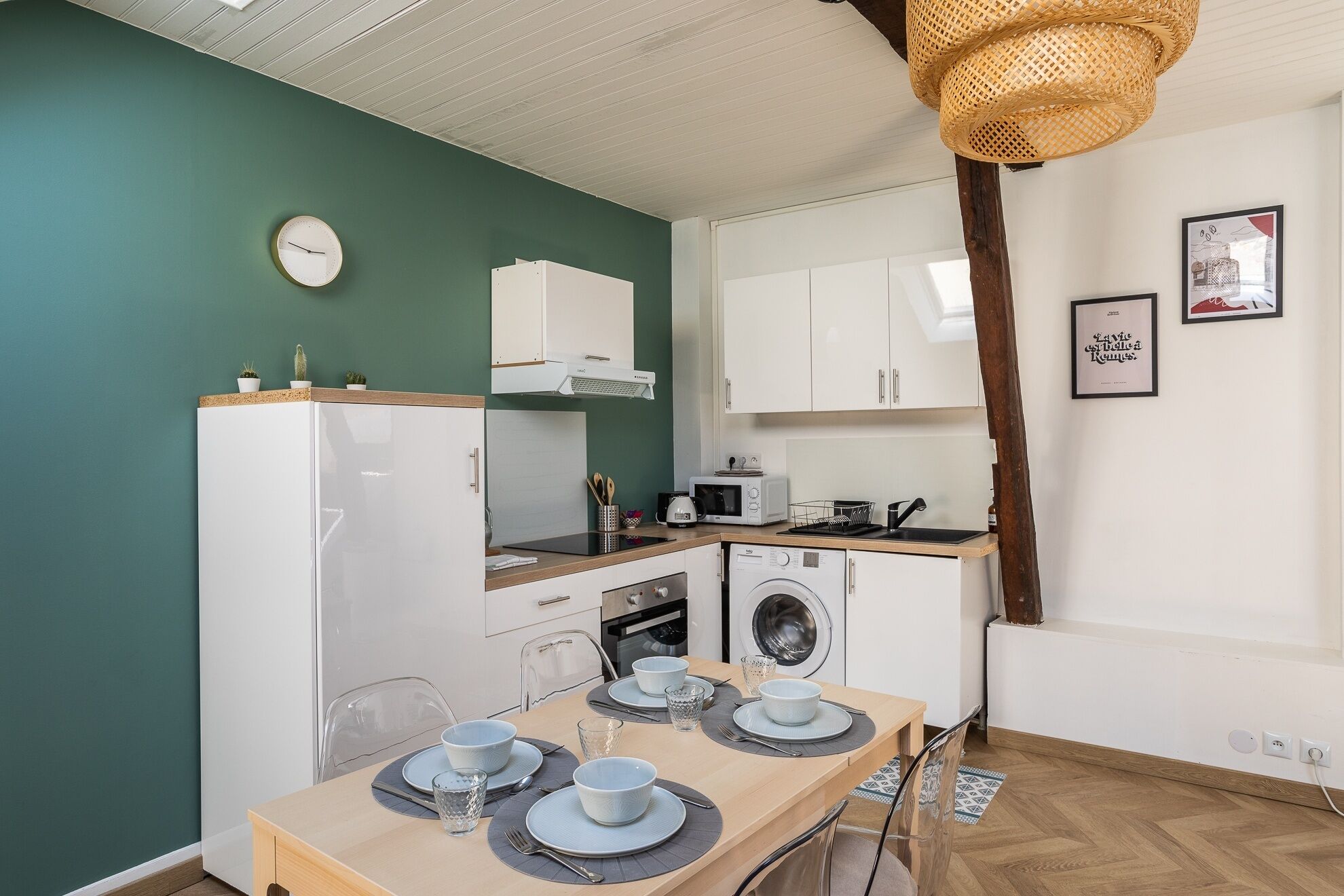 A compact kitchen with a microwave, toaster, and washing machine. There is a dining table set with plates and glasses.