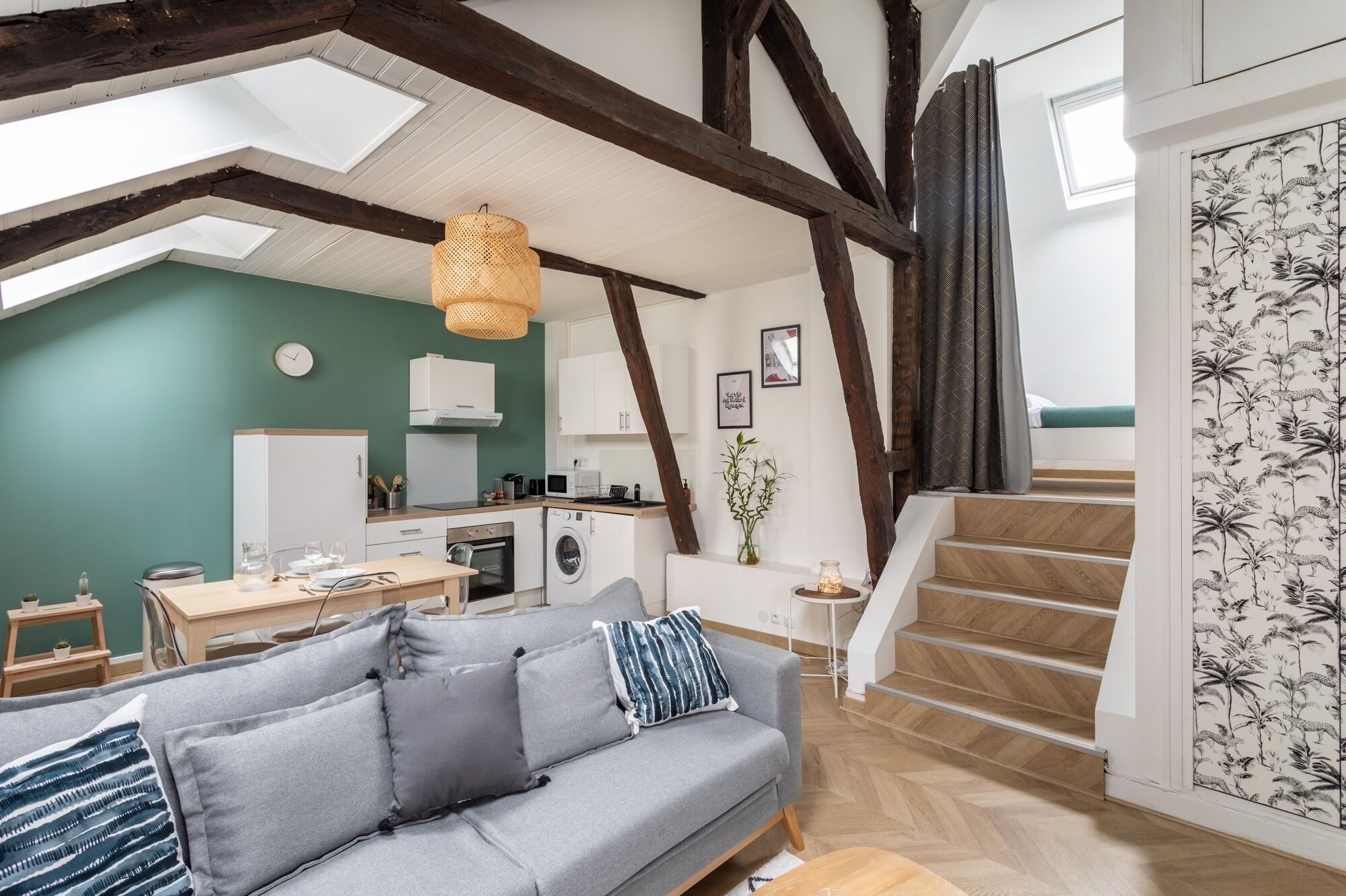 Un espace de vie moderne de style loft, avec des poutres apparentes, un coin cuisine, un espace repas et un escalier menant à un niveau supérieur.