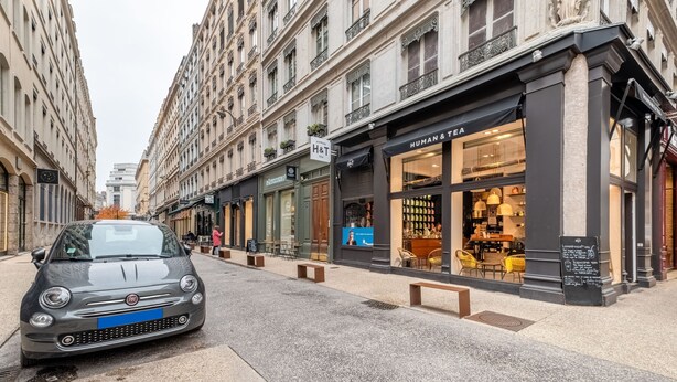 A street view with a car parked on the side, storefronts, and benches along the sidewalk. - Isadora - Ravissant T2 au coeur de Lyon (Lyon)