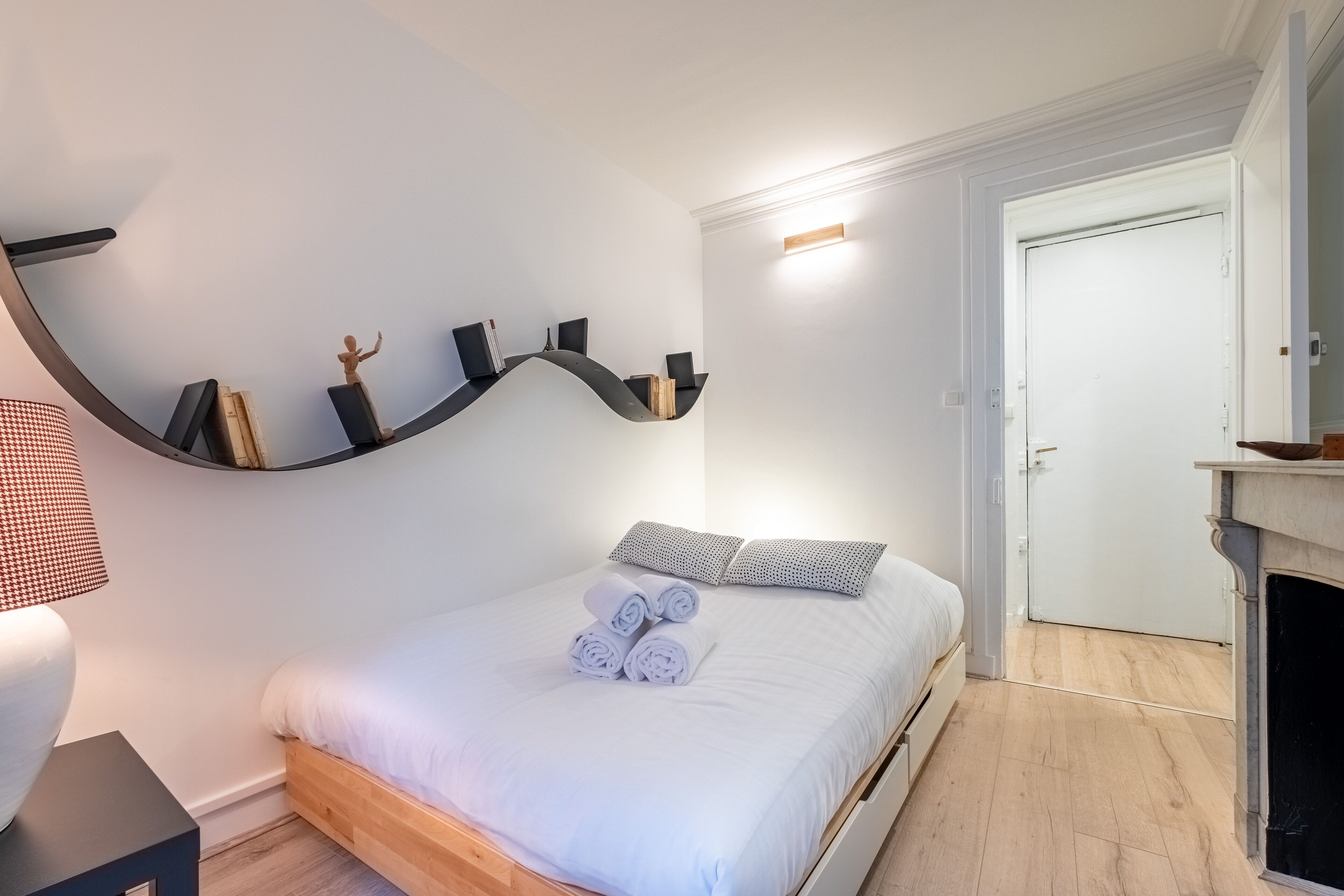 A neatly made bed with rolled towels, a bedside lamp, and a curved shelf with books and a small statue.