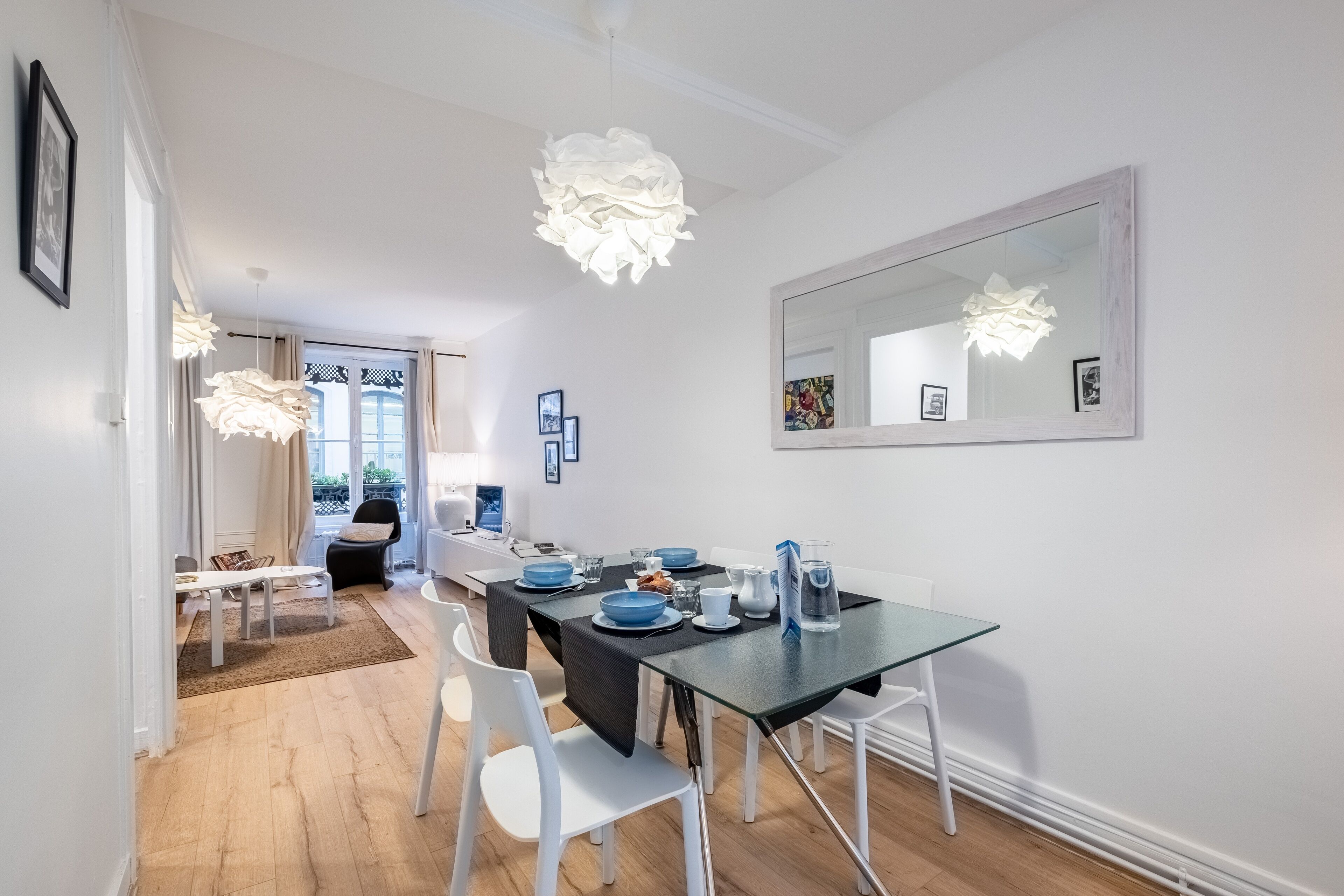A modern dining area with a glass table, white chairs, and a black tablecloth. There are framed pictures on the wall and a large mirror above the table.