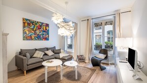 A modern living room with a grey sofa, two white coffee tables, a colorful abstract painting, a unique light fixture, and a view of the outdoors through large windows. - Isadora - Ravissant T2 au coeur de Lyon (Lyon)