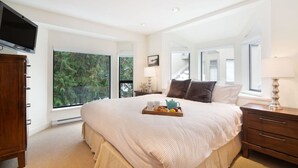 A bedroom with a large bed, a TV mounted on the wall, and a window with a view of greenery. - Prime Location: 4-Bedroom Chalet Style Home (Whistler)