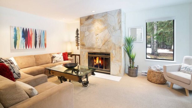 A modern living room with a stone fireplace, a sofa, a coffee table, and a large window with a forest view. - Prime Location: 4-Bedroom Chalet Style Home (Whistler)