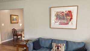 A living room with a blue sofa, a glass coffee table, a patterned rug, a framed abstract painting, and a dining area with a chair and table.
