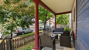 A covered porch with wicker furniture and a table, overlooking a street with parked cars and a fence.