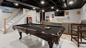 A pool table with balls, a sofa, and a chair in a room with a staircase and arcade machines.