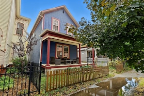 A two-story house with a porch, a wooden fence, and a small garden area. - Nest on Vestry: Spacious Historical Home w/ Jacuzzi & Pool Table (Cleveland)