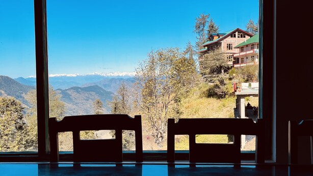 A room with a large window offering a view of a mountainous landscape and a building with a red roof. - An artsy and peaceful mountain stay located in an offbeat village of Shoja (Banjar)