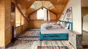 A cozy attic bedroom with wooden walls, a sloped ceiling, a bed with a blue headboard, and a small window with curtains. - An artsy and peaceful mountain stay located in an offbeat village of Shoja (Banjar)