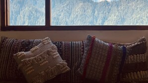 A view of a mountain range through a window, with a sofa and patterned cushions in the foreground. - An artsy and peaceful mountain stay located in an offbeat village of Shoja (Banjar)