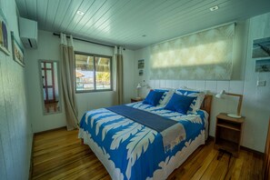 A bedroom with a bed, wooden flooring, a window with curtains, a nightstand with a lamp, and wall decorations. - Gorgeous Over Water Bungalow in Bora Bora. Perfect spot Honeymooners & Families (Bora-Bora)
