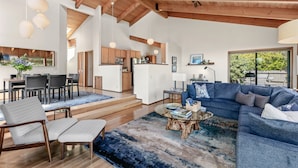 A spacious living room with a large blue sectional sofa, a glass coffee table, and a dining area with a wooden table and chairs.