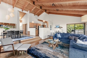 A spacious living room with a large blue sectional sofa, a glass coffee table, and a dining area with a wooden table and chairs.