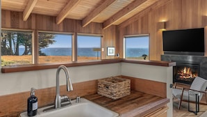 A modern kitchen with wooden beams, large windows with ocean views, a fireplace, and a stone countertop.