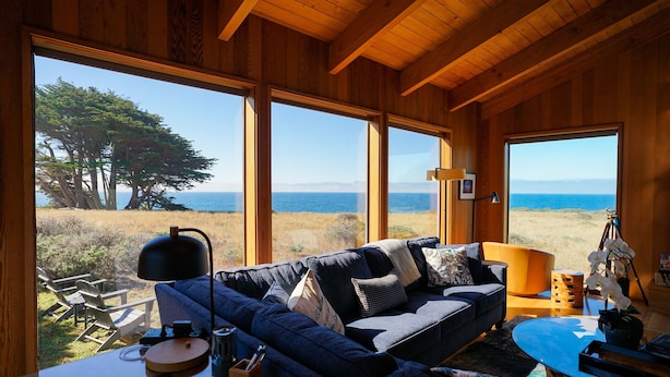 A cozy room with a large window overlooking a beach and ocean, furnished with a sofa, chairs, and a coffee table.