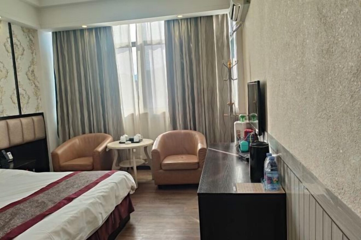 A hotel room with a bed, two armchairs, a small round table, a desk with a computer and a bottle of water, and a TV on the wall.