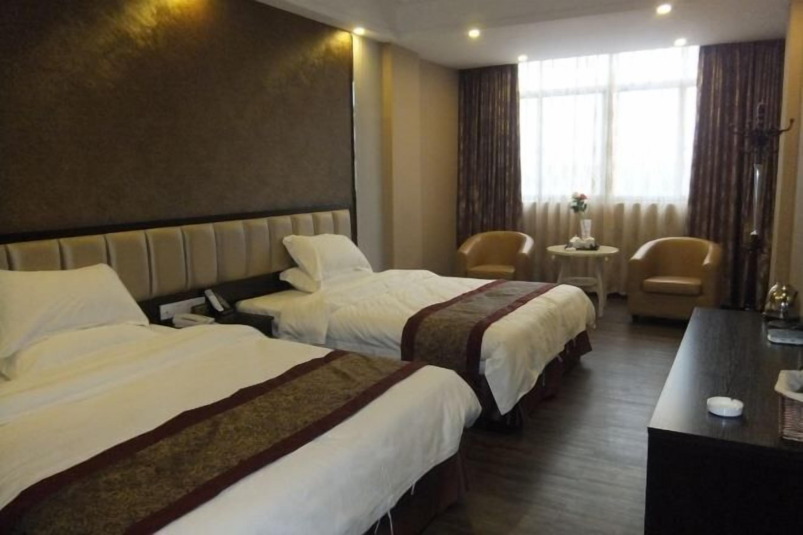 A hotel room with two beds, a desk, two chairs, a small table with a vase, and a window with curtains.