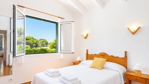 A bedroom with a large bed, a window with an open curtain, a bedside table with a lamp, and a view of greenery outside.