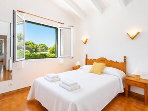 Un dormitorio con una cama grande, una ventana con cortina abierta, una mesita de noche con lámpara y vistas al exterior con vegetación.