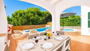 An outdoor dining area with a pool and greenery in the background.