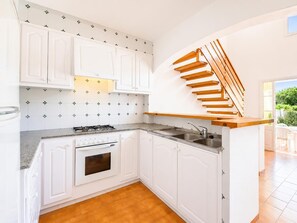 Una cocina con armarios blancos, estufa a gas, horno empotrado, una escalera de madera y vistas al exterior con vegetación.