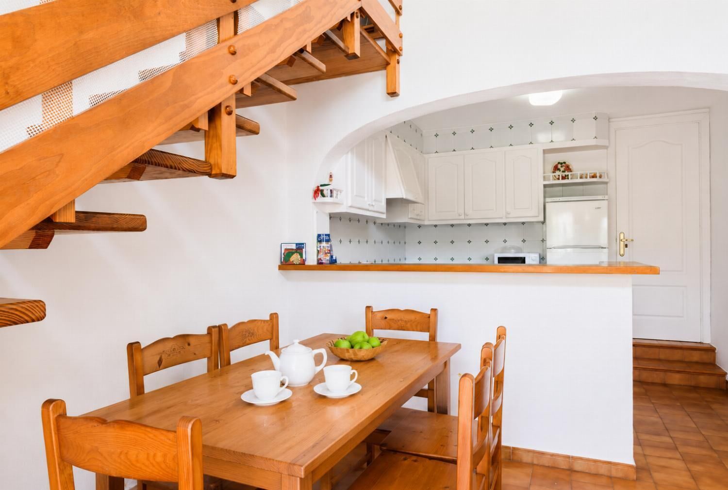 Une table à manger en bois avec des chaises, une cuisine avec des meubles blancs et un escalier.